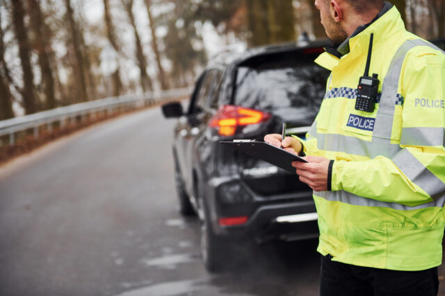 Male police officer in green uniform standing with notepad near car.